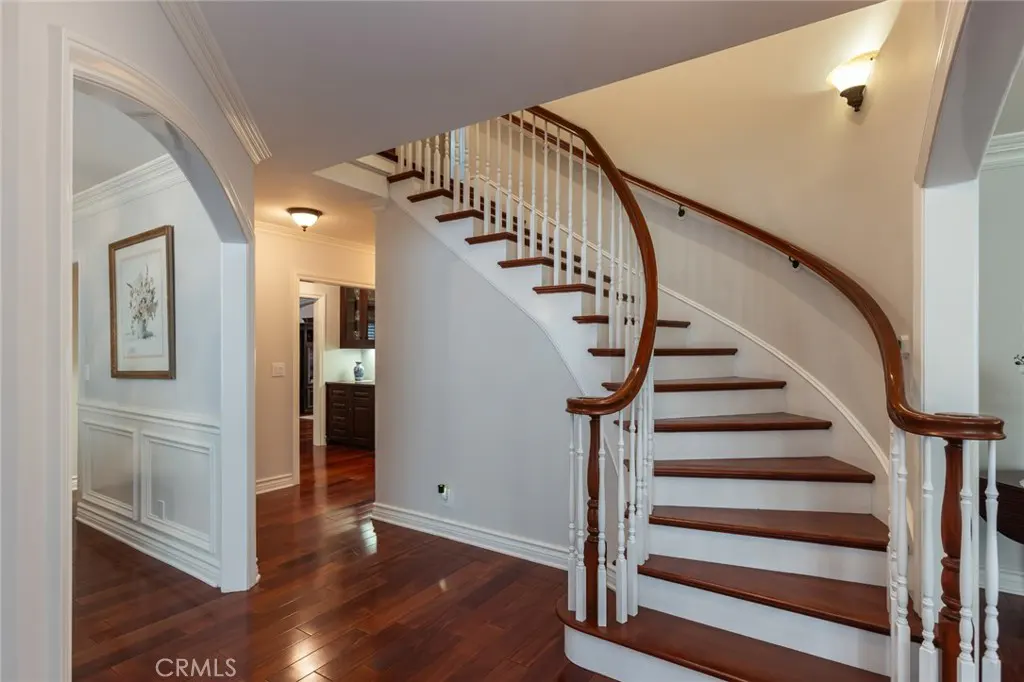 A foyer with a curved staircase, white walls, and dark wood floors. An arched doorway leads to another room.