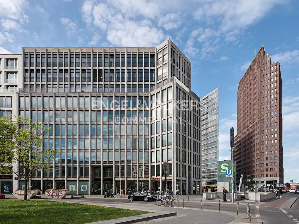 Modern office building with a grid facade next to a tall brown skyscraper and a subway entrance under a blue sky.