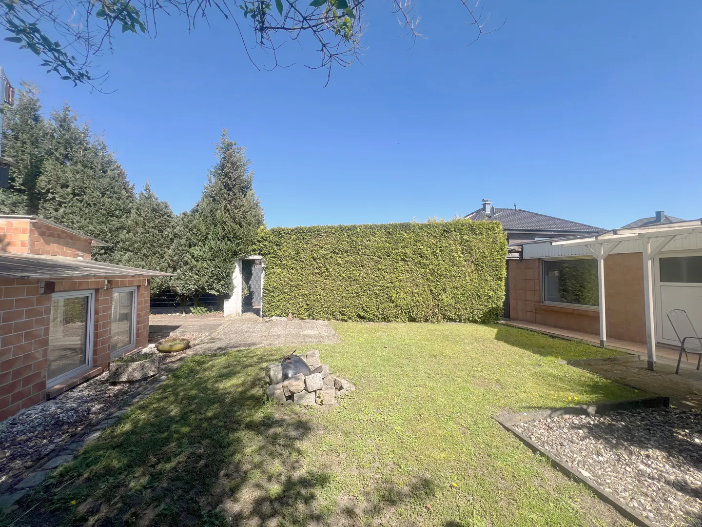 A backyard with green grass, a tall hedge, a brick building, and a stone patio on a sunny day.