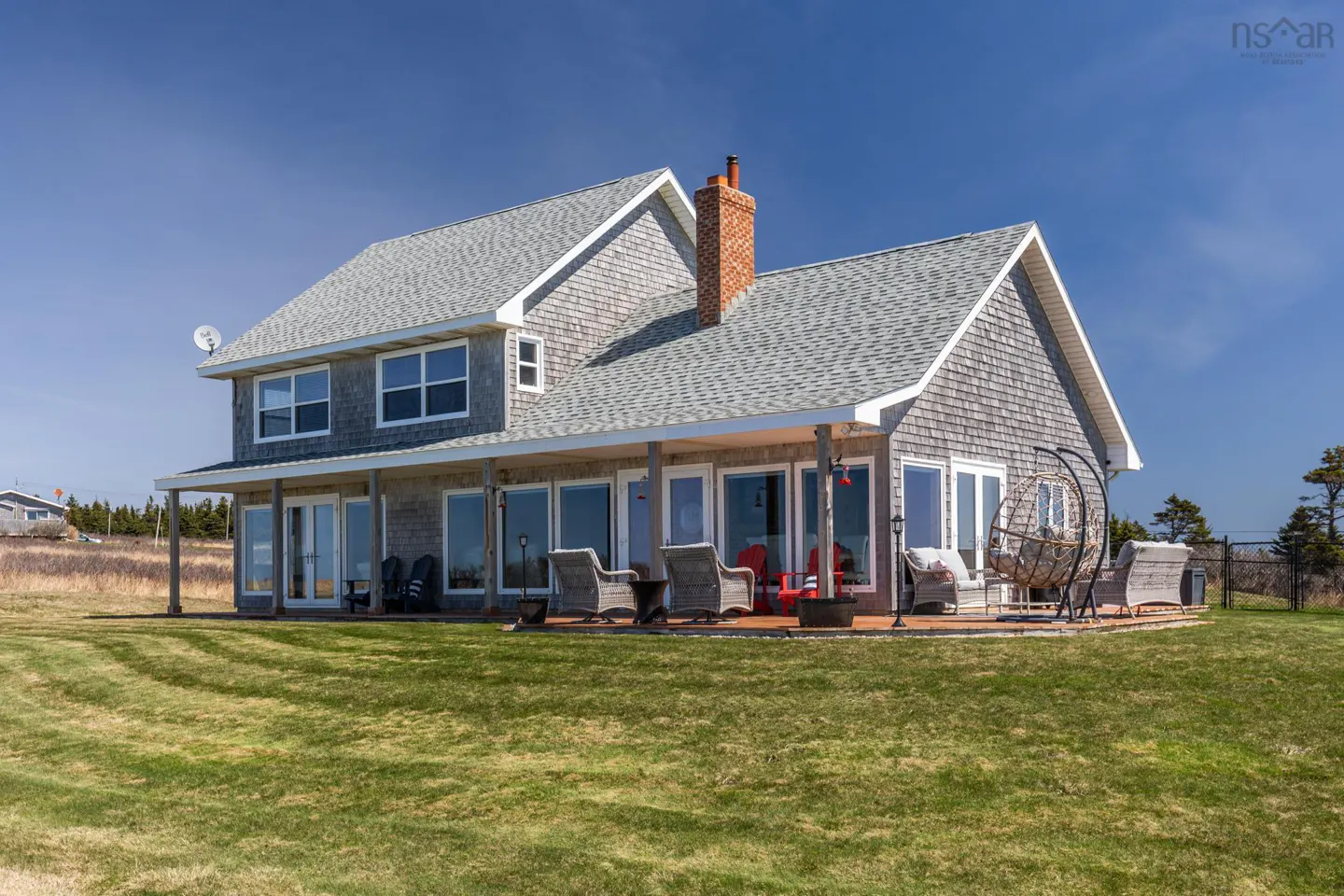 Two-story house with gray shingles, white trim, and a brick chimney. A porch with chairs and a hanging swing faces a green lawn.