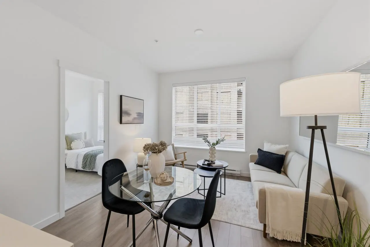 Bright, modern apartment living room with a glass table, black chairs, and a cream-colored sofa. A bedroom is visible through a doorway.