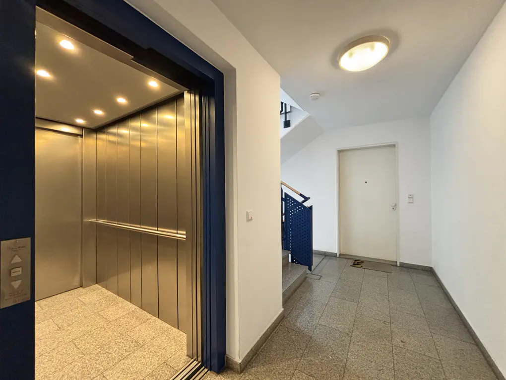Hallway view with an open elevator with a blue frame, stainless steel interior, and a tiled floor. A white door and stairs are visible in the background.