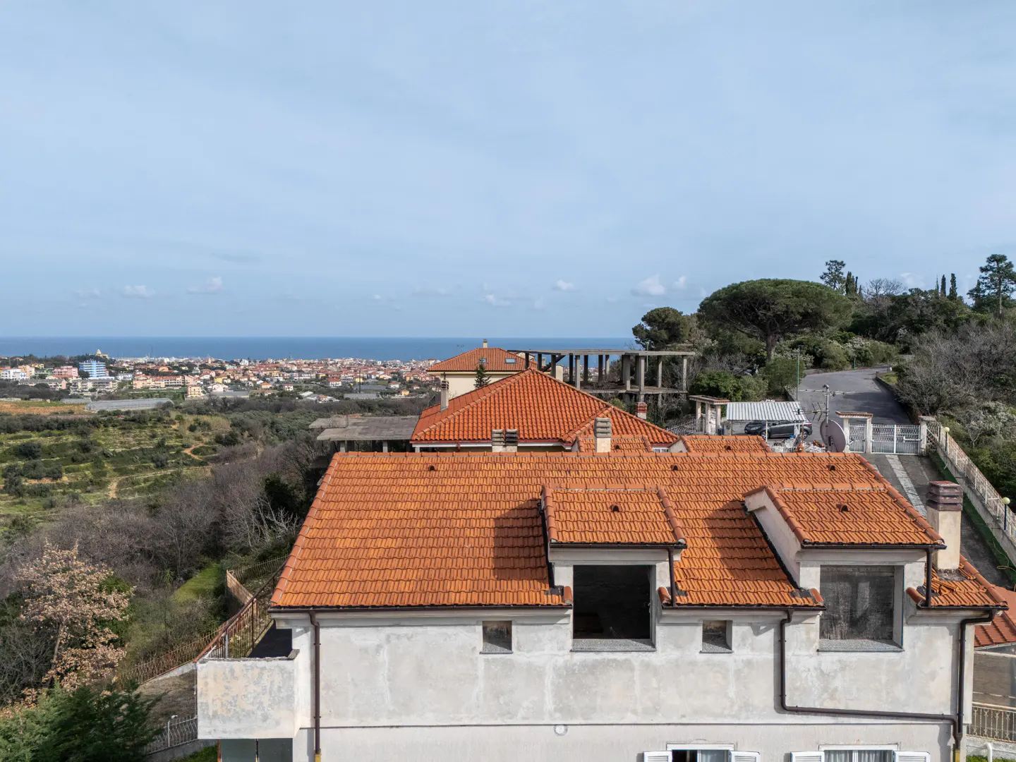 High-angle view of a house with a terracotta tile roof overlooking a coastal town and the sea under a clear blue sky.