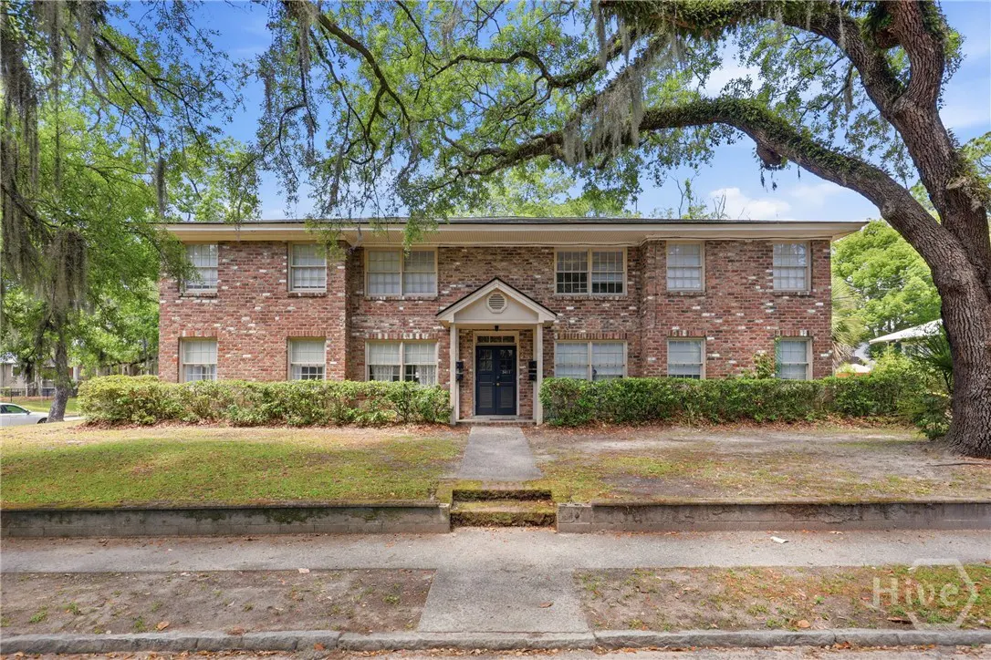 Two-story brick apartment building with a black door and green bushes, under a large tree with Spanish moss.