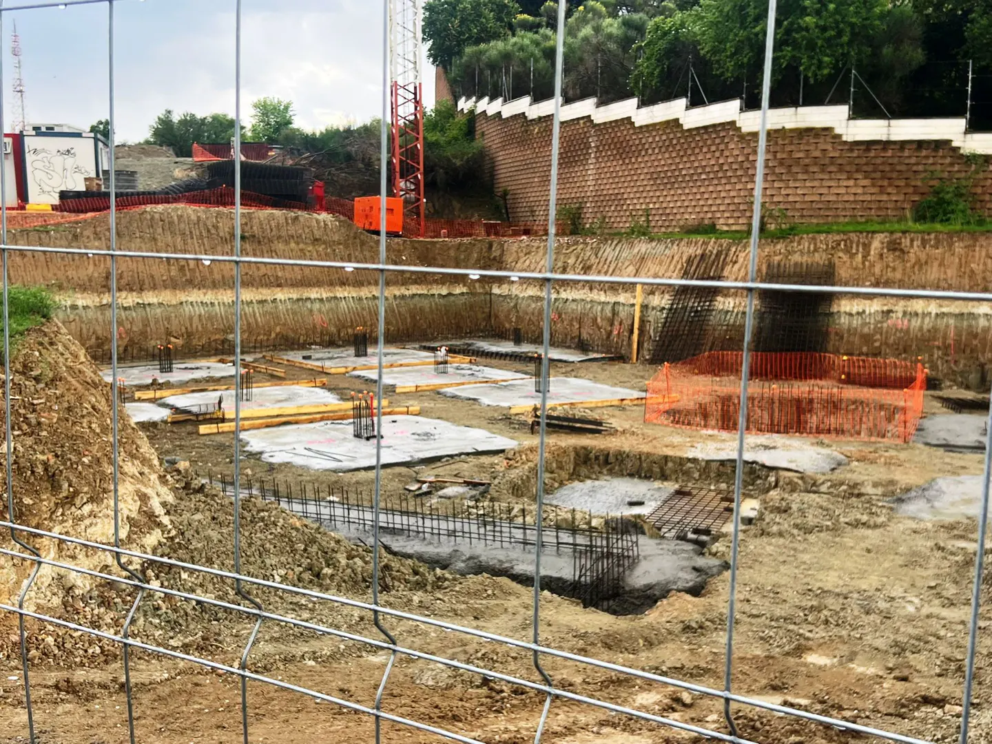 Construction site with a large pit, concrete foundations, rebar, and an orange crane visible behind a wire fence.