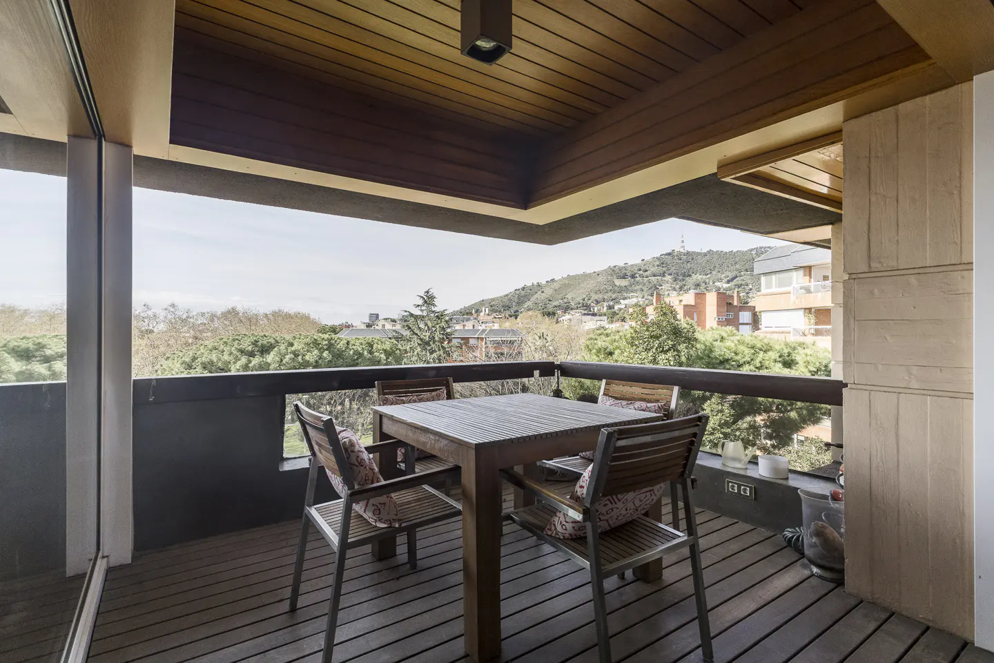 A wood-decked balcony with a table and four chairs overlooks trees and a distant hill. The ceiling is wood, and a light fixture is visible.