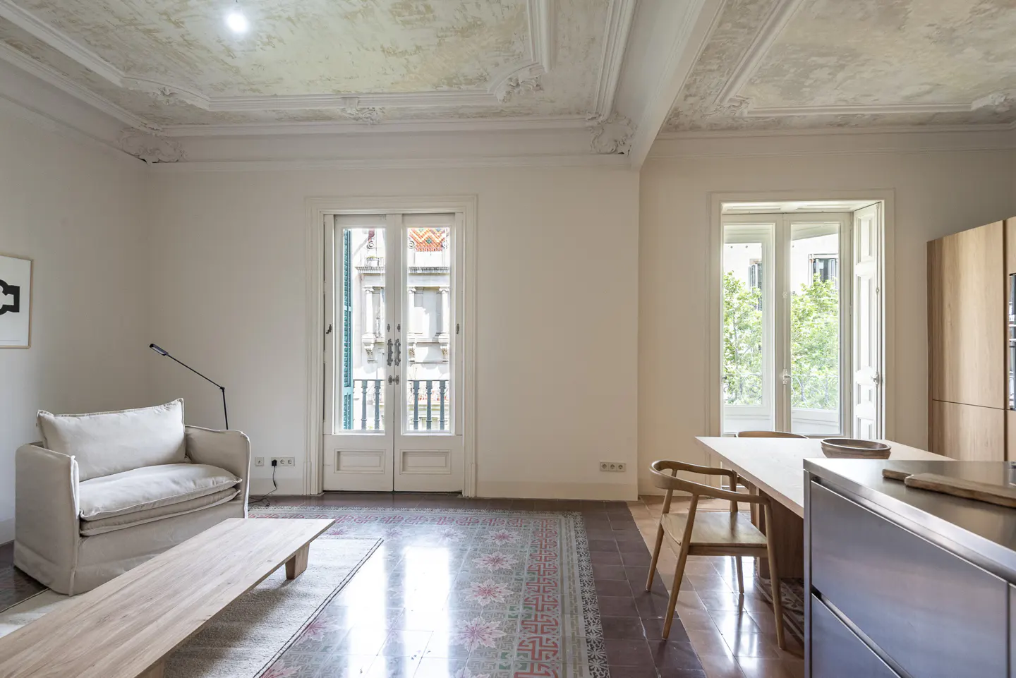 Bright, airy living space with white walls, ornate ceiling, and patterned tile floor. A white armchair, wood table, and kitchen area are visible.