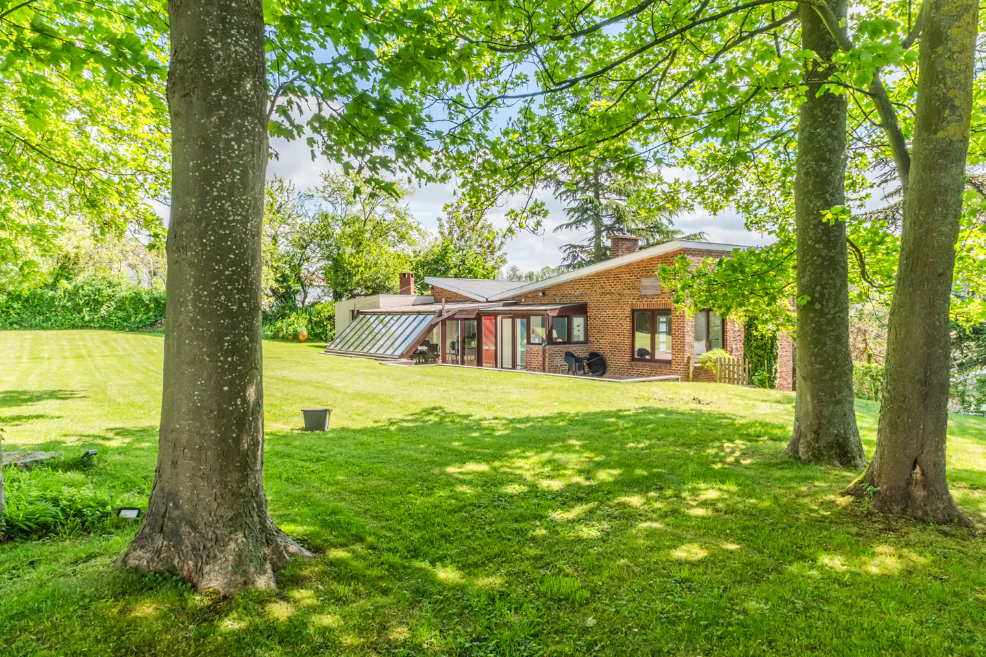 Exterior view of a brick house with a glass sunroom, seen through trees on a green lawn.