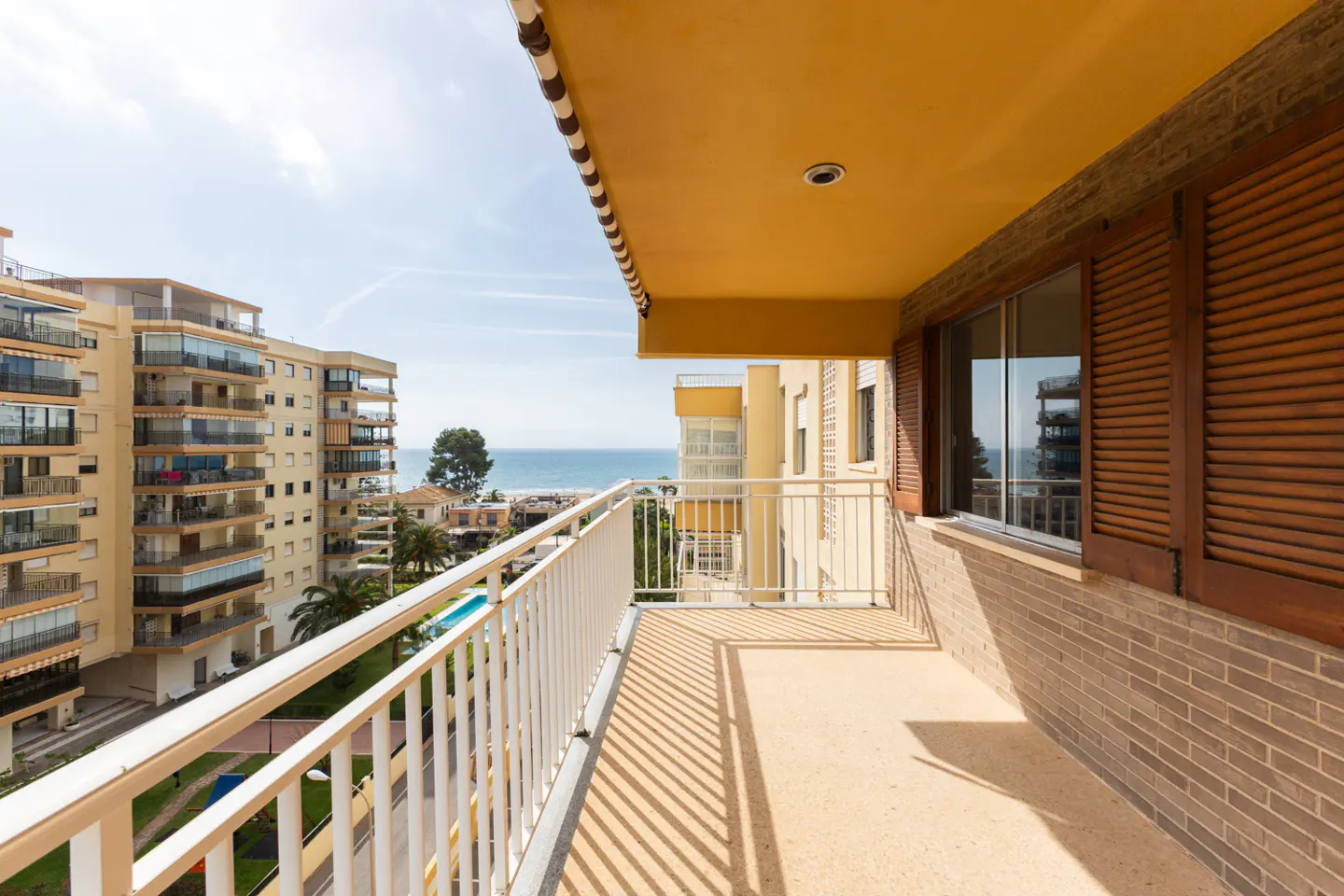Balcony view of a tan building and the ocean. White railings cast shadows on the floor. Brown shutters are on the window.