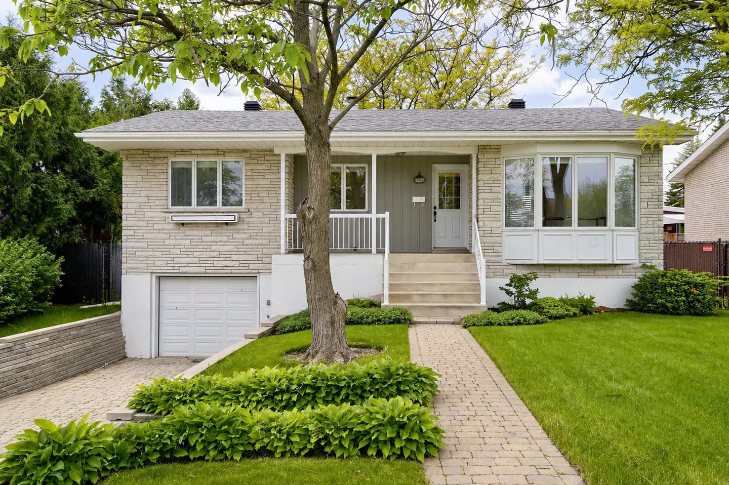 A one-story house with a gray brick facade, white trim, and a gray roof. A tree stands in the front yard, with a brick walkway leading to the front door.