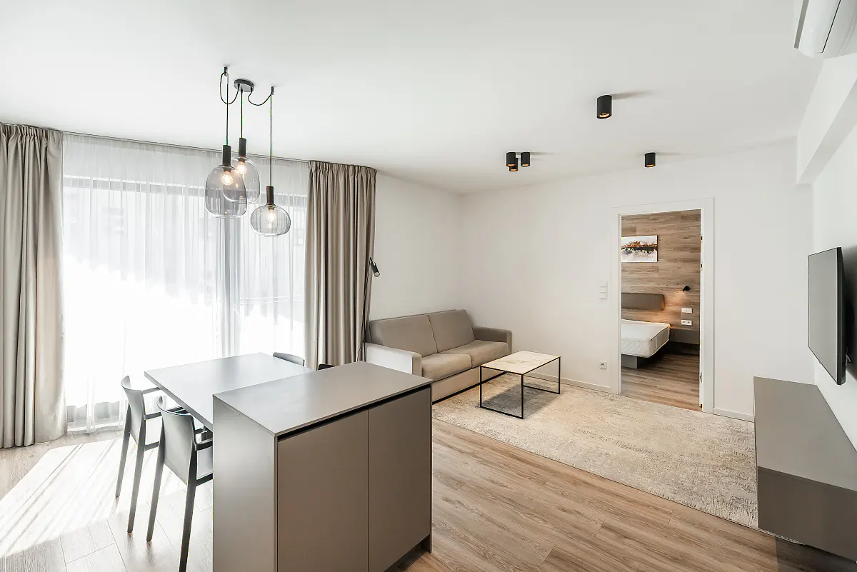 Modern apartment living area with a dining table, grey sofa, coffee table, and a view into a bedroom with wood accents.