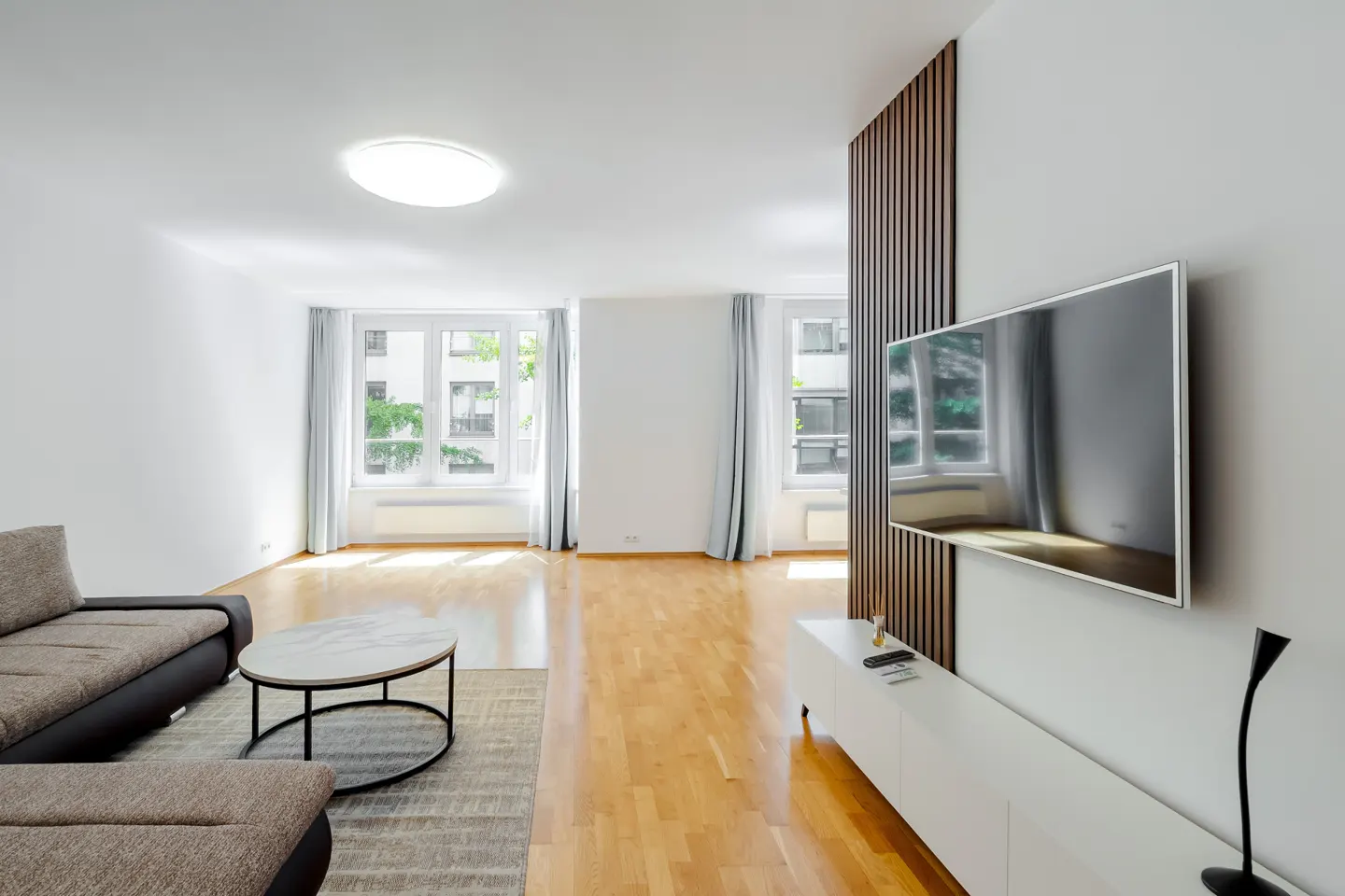 Bright living room with wood floors, white walls, and large windows. A gray sofa, round table, and TV with wood accent wall are featured.