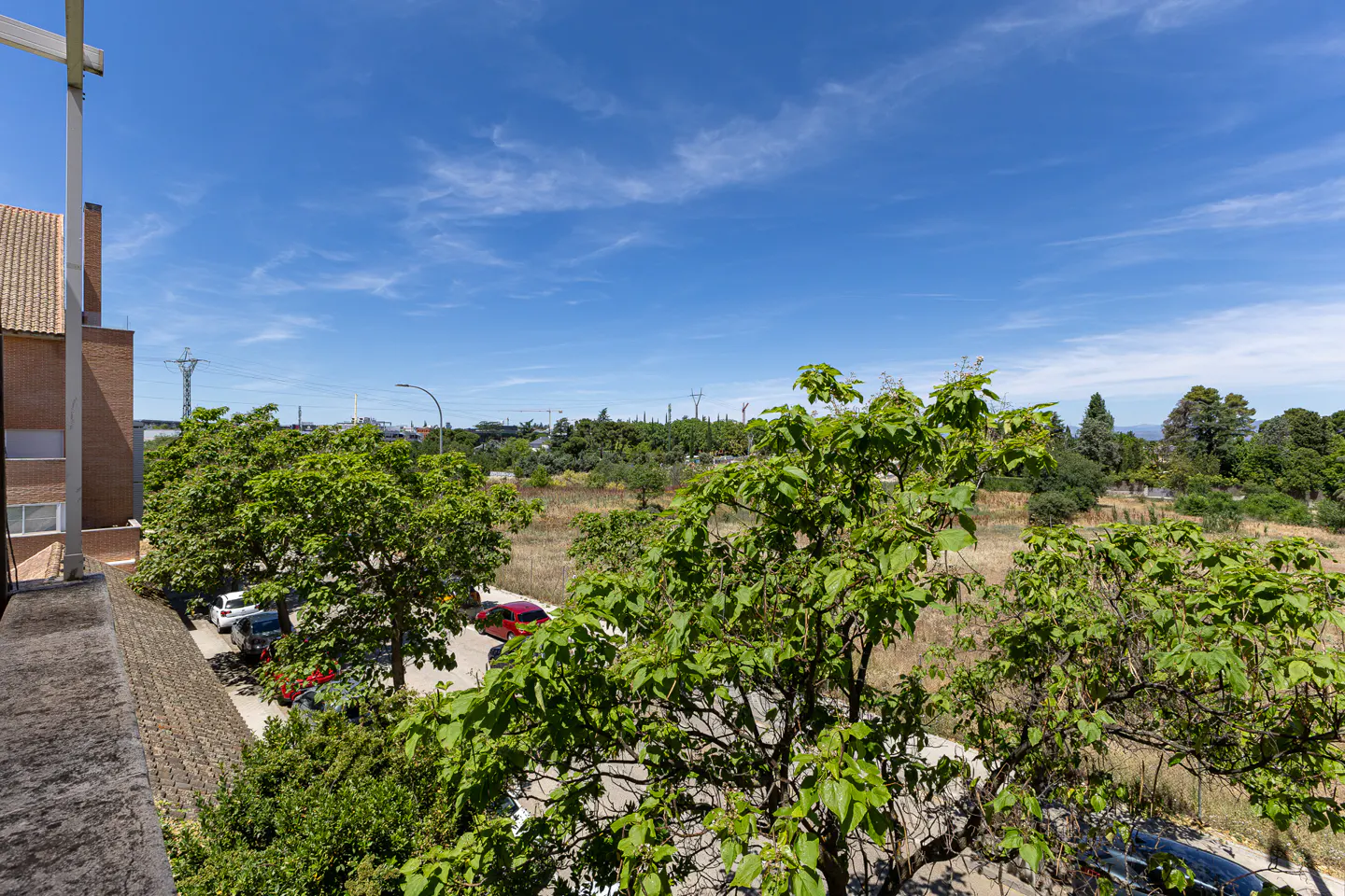 Elevated view of a parking area with trees, a dry field, and distant buildings under a bright blue sky.