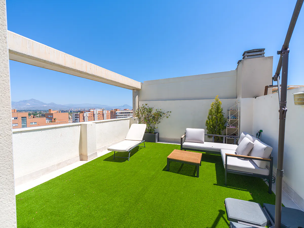 Rooftop terrace with artificial grass, lounge seating, a sun lounger, and a city view under a clear blue sky.