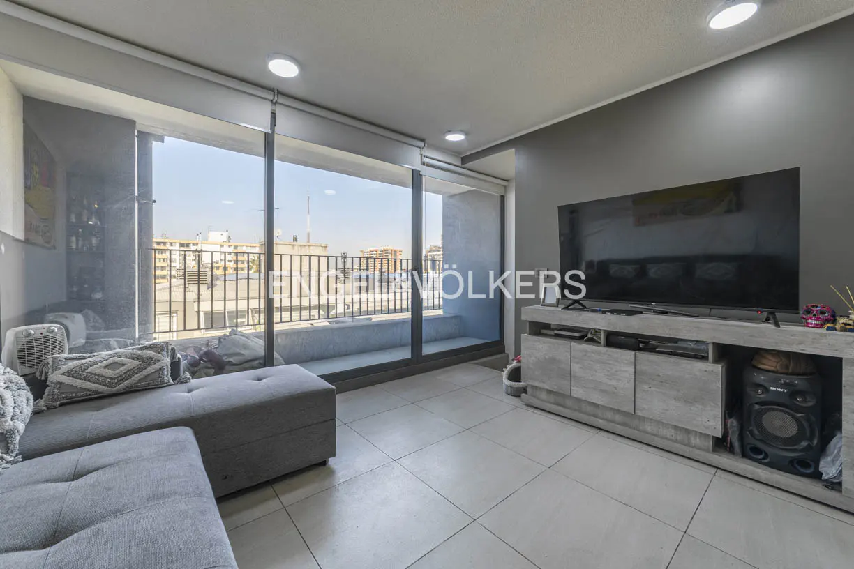 Living room with gray sofa, TV, and balcony overlooking city. Engel & Völkers logo on the window.