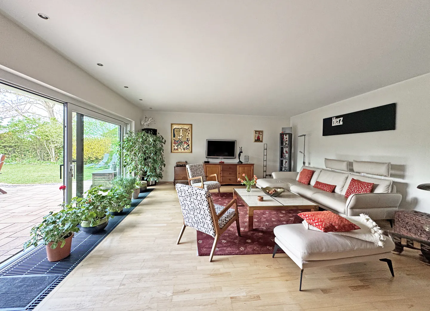Bright living room with light wood floors, a white sectional sofa with red pillows, and a large sliding glass door to a green yard.
