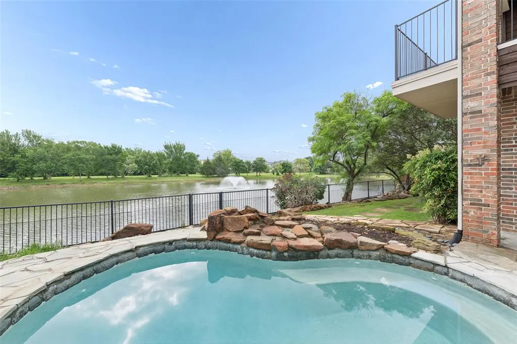 Backyard view of a pool with a stone border, a lake with a fountain, and a balcony on a brick house.