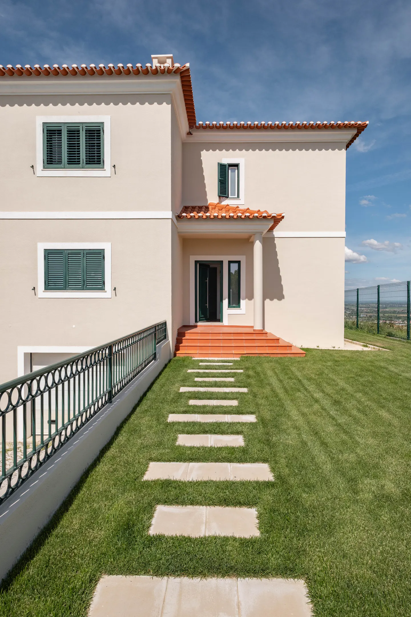 Two-story house with beige walls, red tile roof, and green shutters. Stone path leads to the front door on a green lawn.