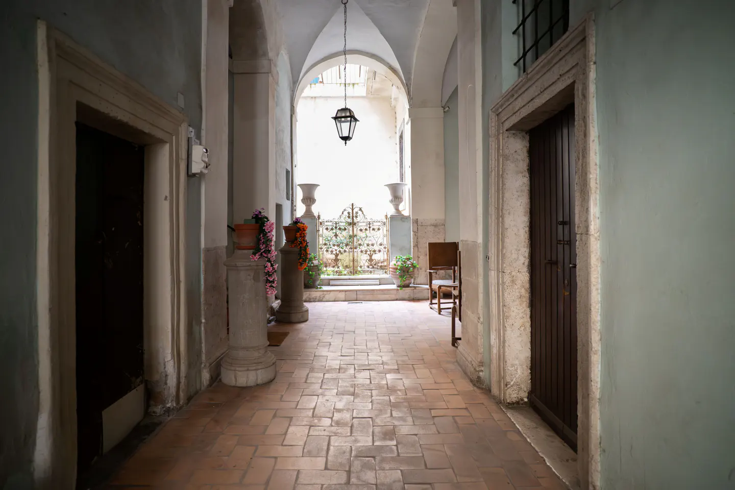 Arched hallway with brick floor, leading to a courtyard with plants and a decorative gate. A hanging lantern is centered in the archway.