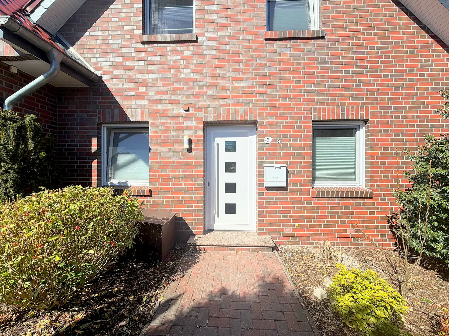 Red brick house with a white front door, mailbox, and three windows. A brick walkway leads to the entrance, with bushes on either side.