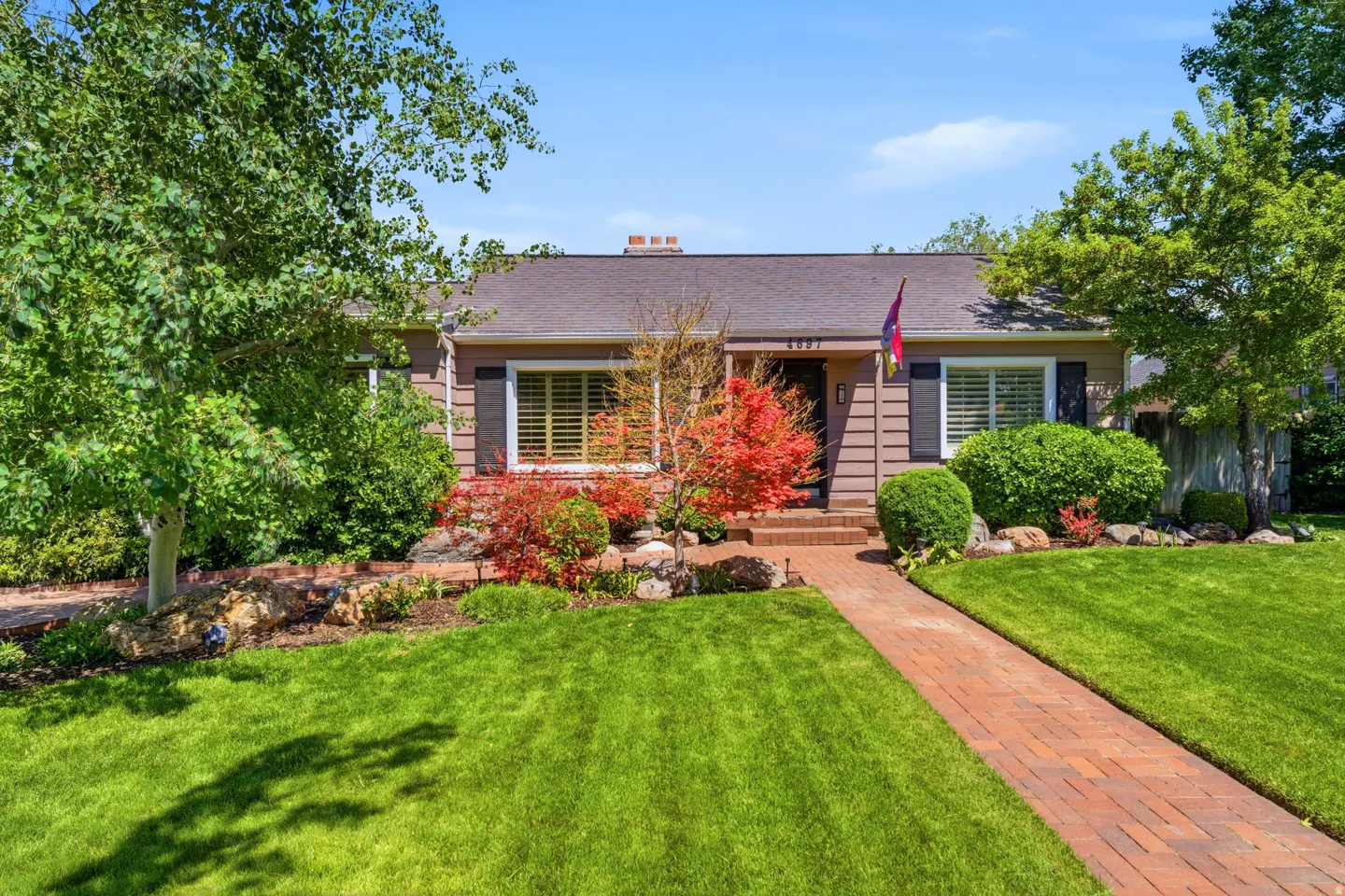 A single-story brown house with a brick walkway, green lawn, and trees under a blue sky.