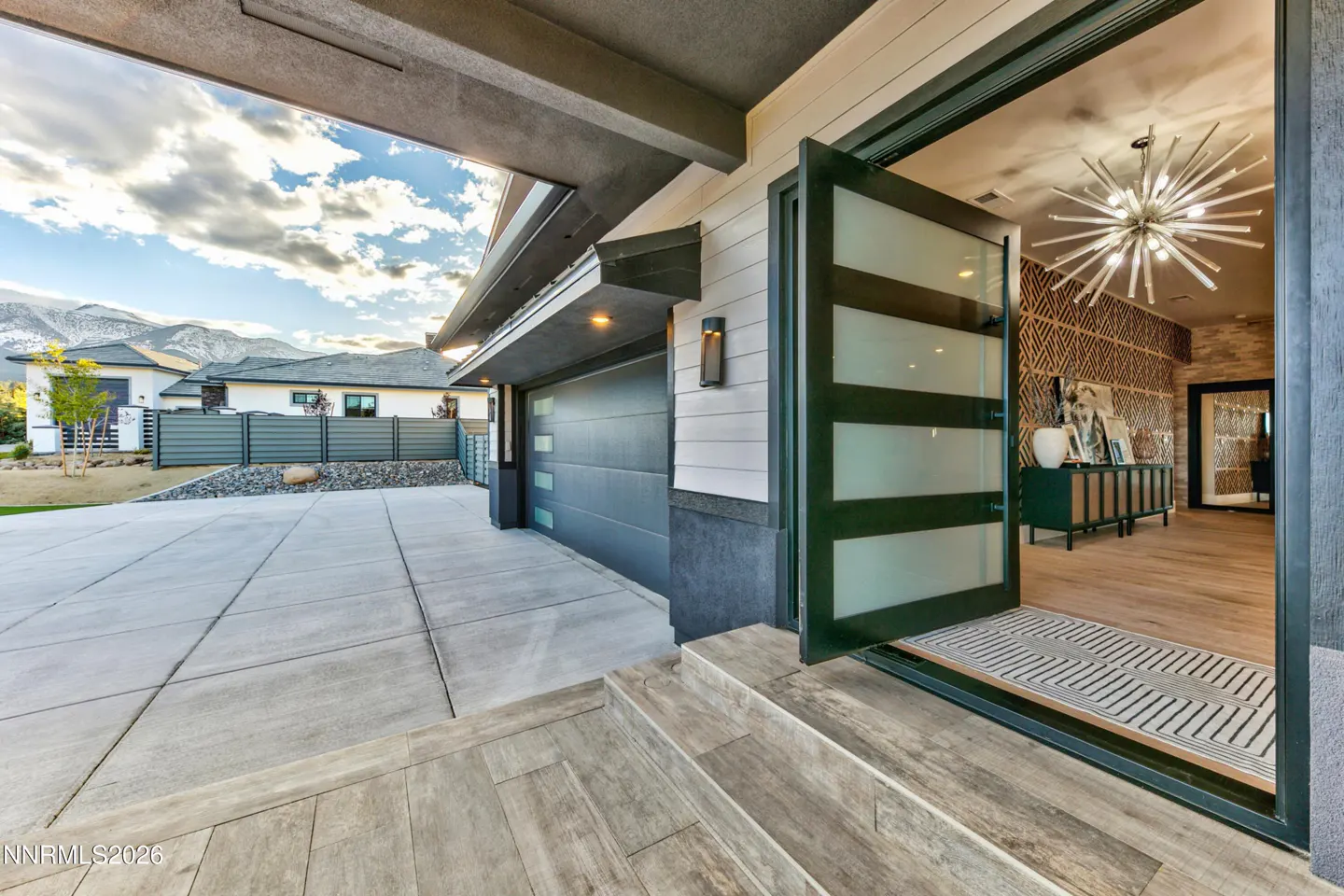 Open frosted glass door reveals a modern home interior with a starburst chandelier, console table, and wood floors. Garage and driveway visible.