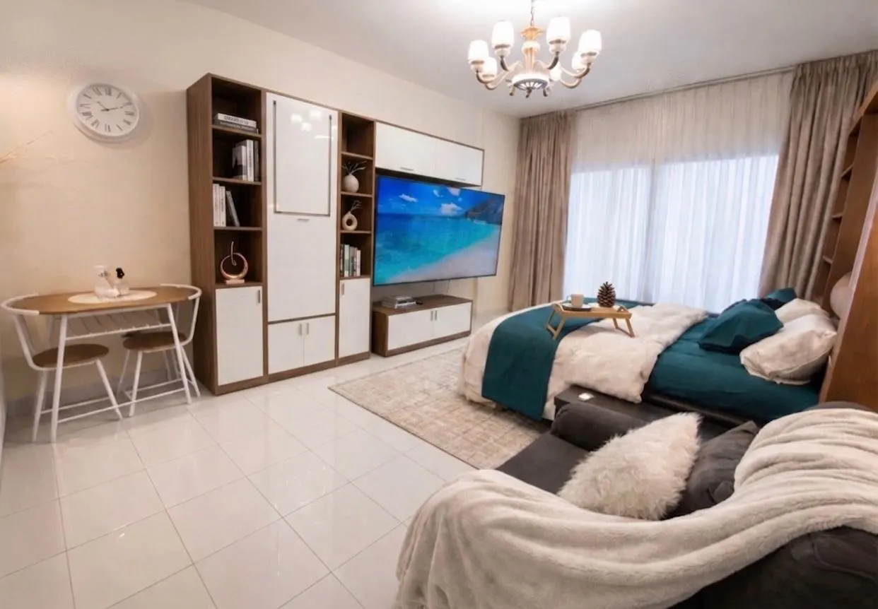 Bright studio apartment with a bed, TV, dining table, and built-in storage. The bed has teal accents and a tray with a mug.