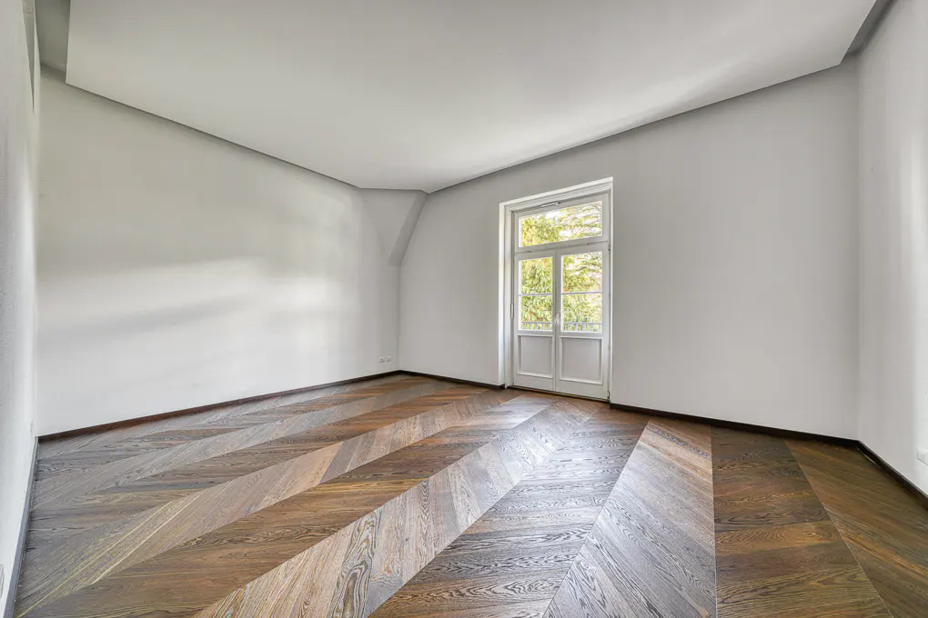 Empty room with white walls, a high angled ceiling, and dark wood chevron-patterned flooring leading to a glass door.