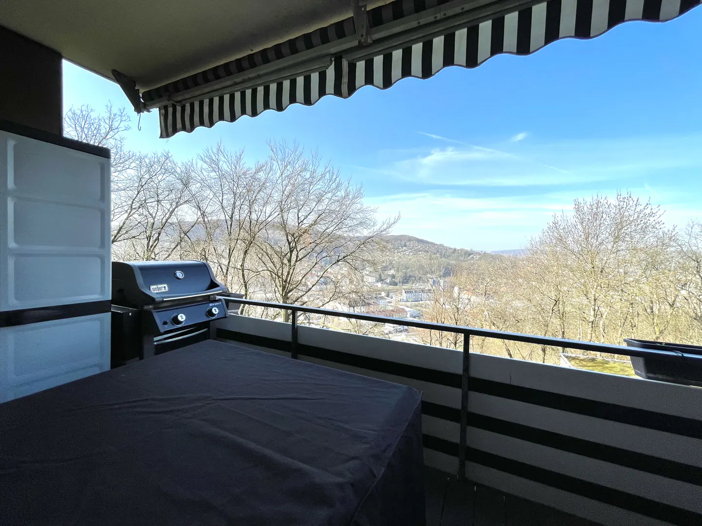 Balcony view with a black grill, covered table, and storage unit. A black and white awning provides shade, overlooking a scenic landscape.