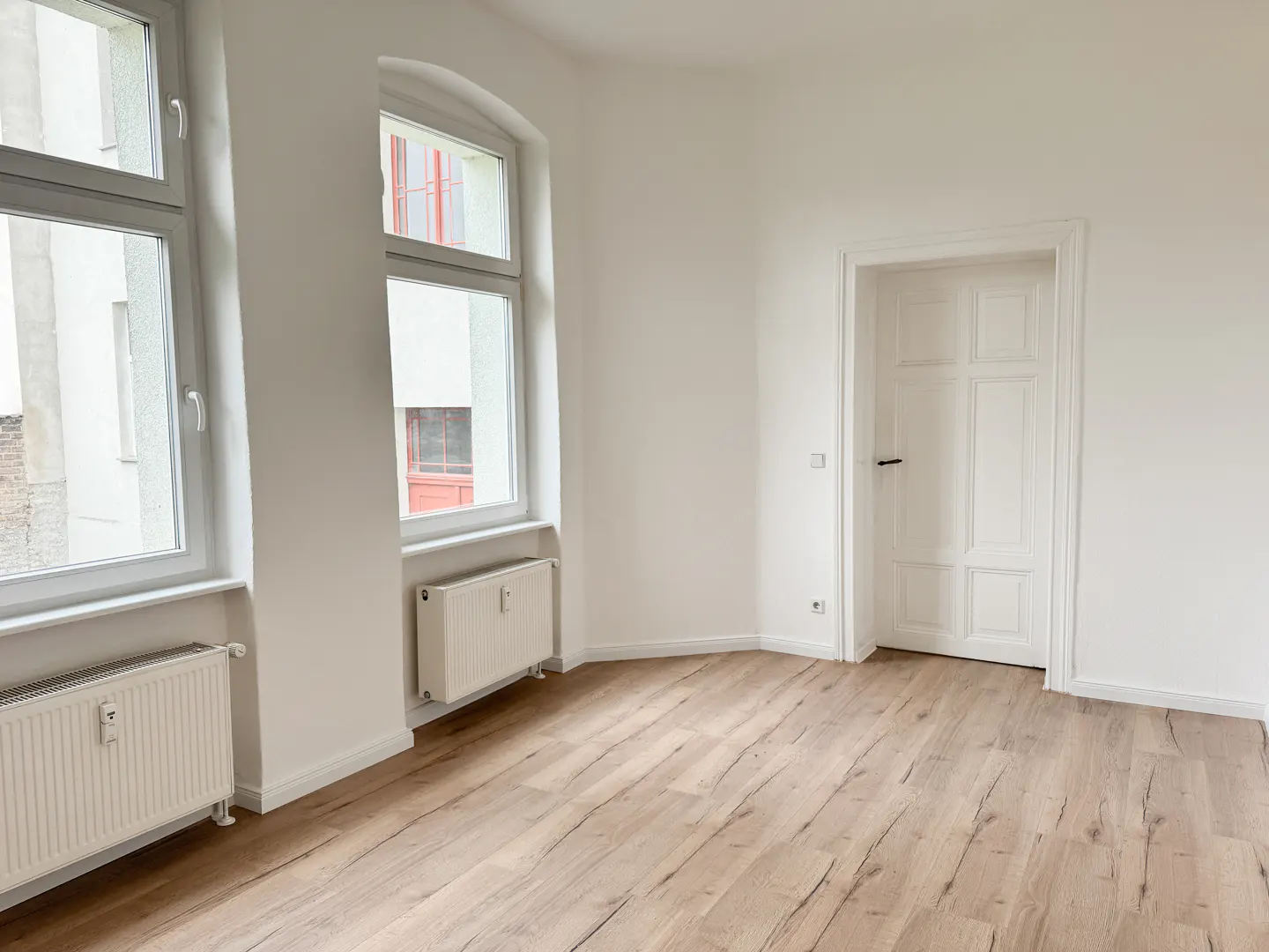 Bright, empty room with light wood floors, white walls, two windows, and a white door. Radiators are under the windows.
