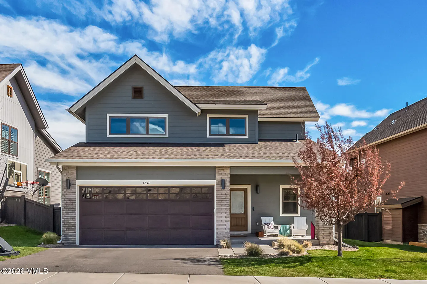 Two-story gray house with brown roof, garage, and stone accents. Two white chairs sit on the porch. Blue sky with clouds above.