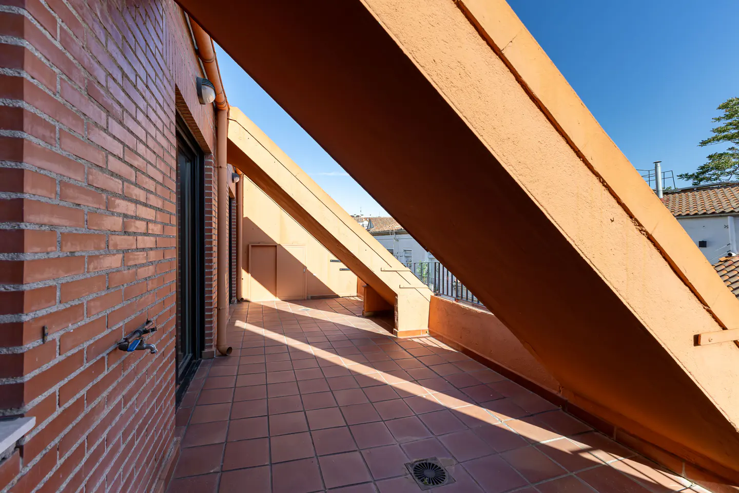 Exterior view of a balcony with orange beams, brick wall, and tiled floor under a blue sky.