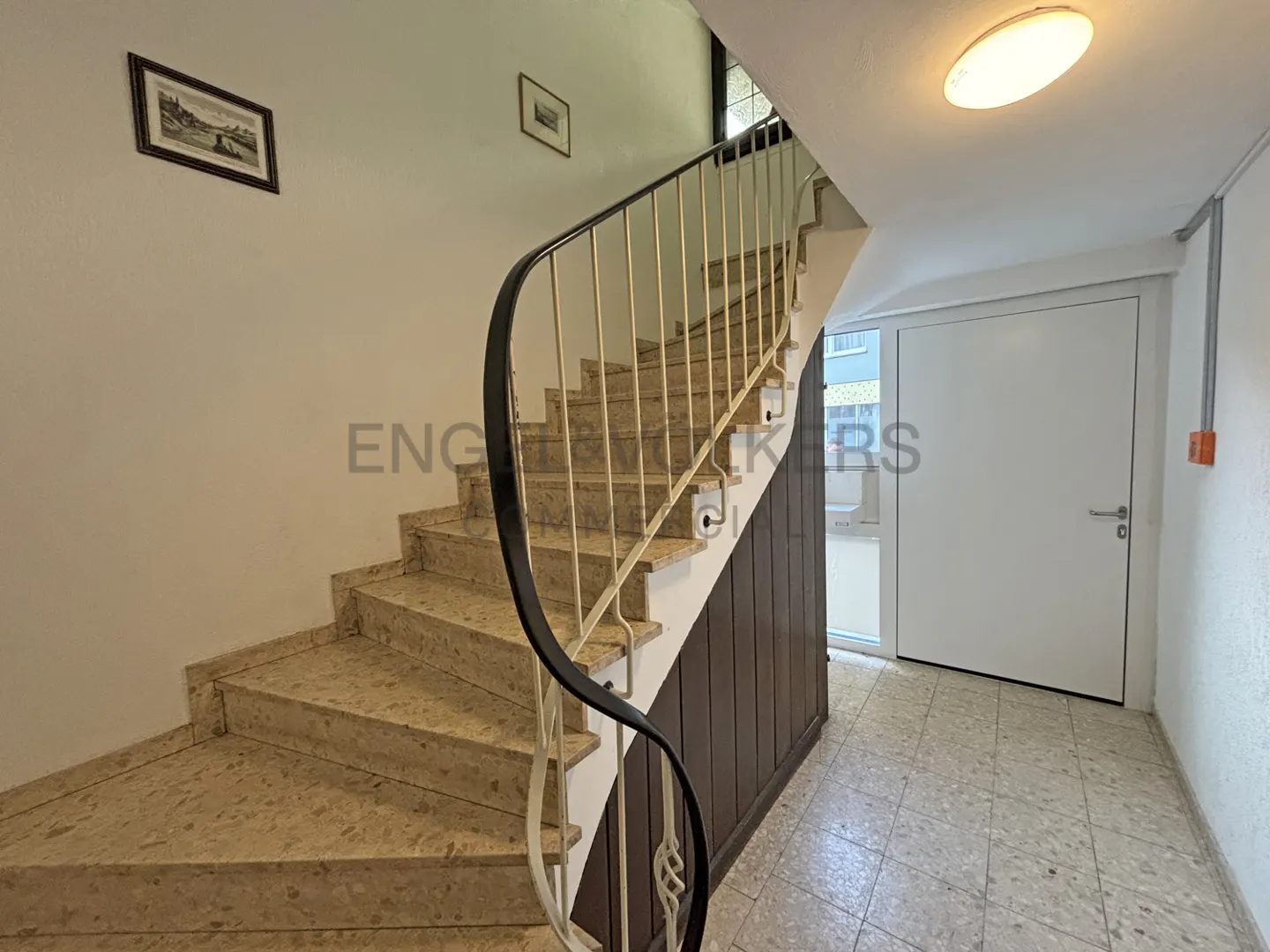Interior view of a staircase with beige stone steps, a black handrail, and a white wall with framed pictures. A white door is visible on the right.