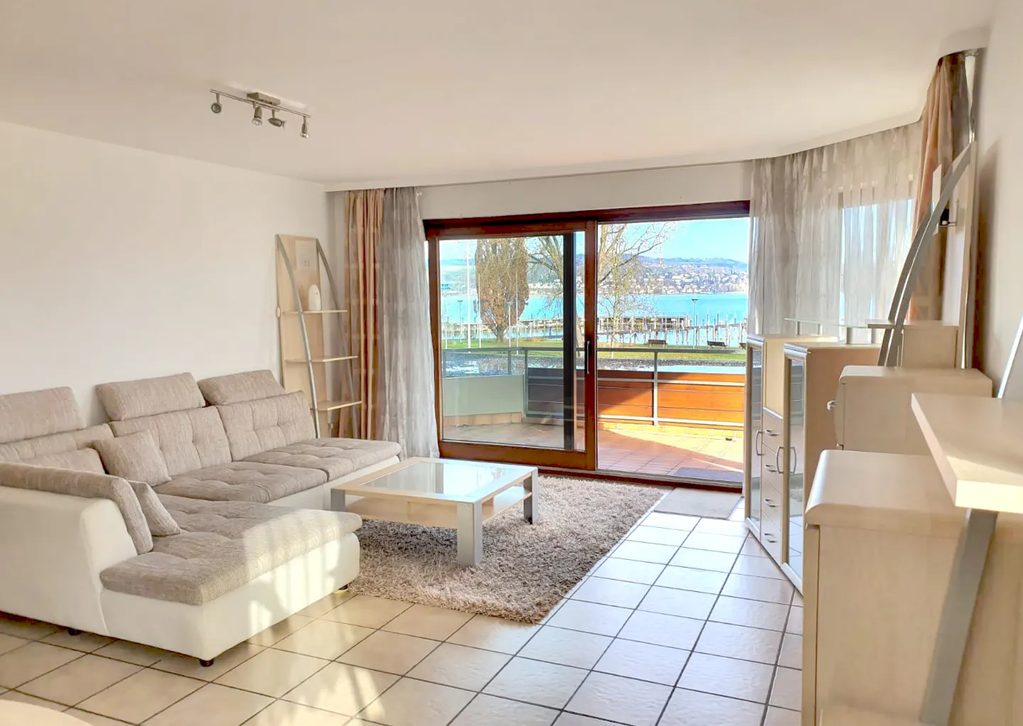 Bright living room with a beige sectional sofa, a glass coffee table on a rug, and a sliding glass door to a balcony with a lake view.