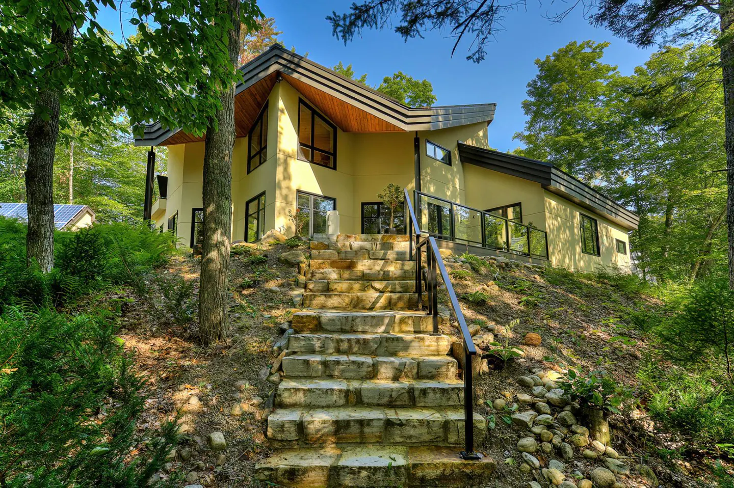 Exterior view of a modern, yellow house with a dark roof, stone steps, and lush green trees.