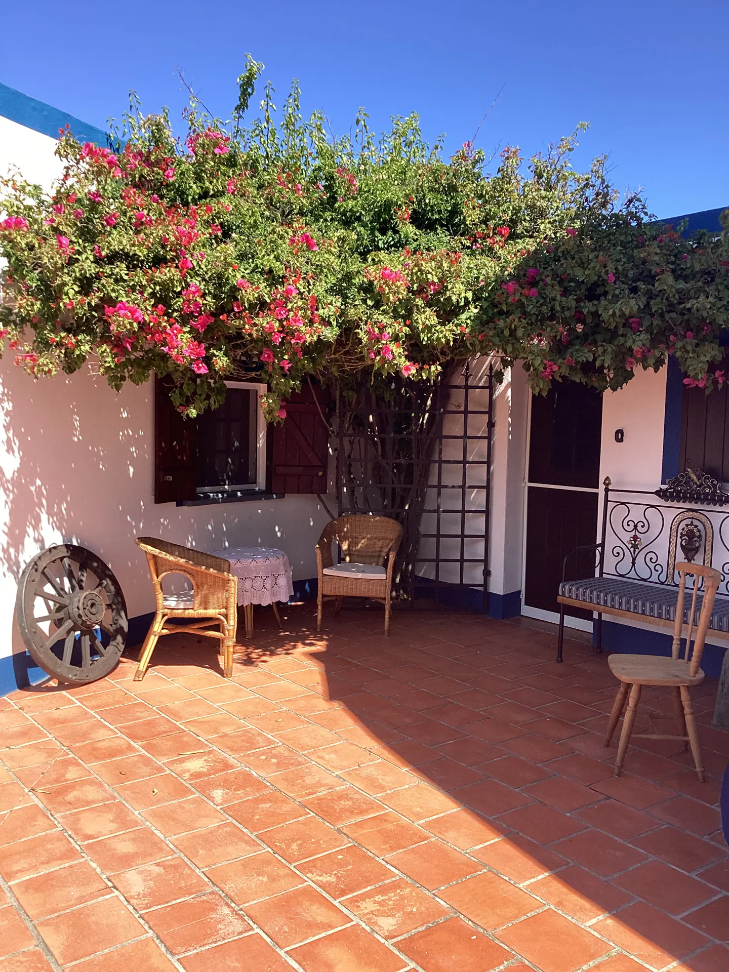 Outdoor patio with terracotta tiles, white walls, and bougainvillea. Wicker chairs, a bench, and an old wooden wheel decorate the space.