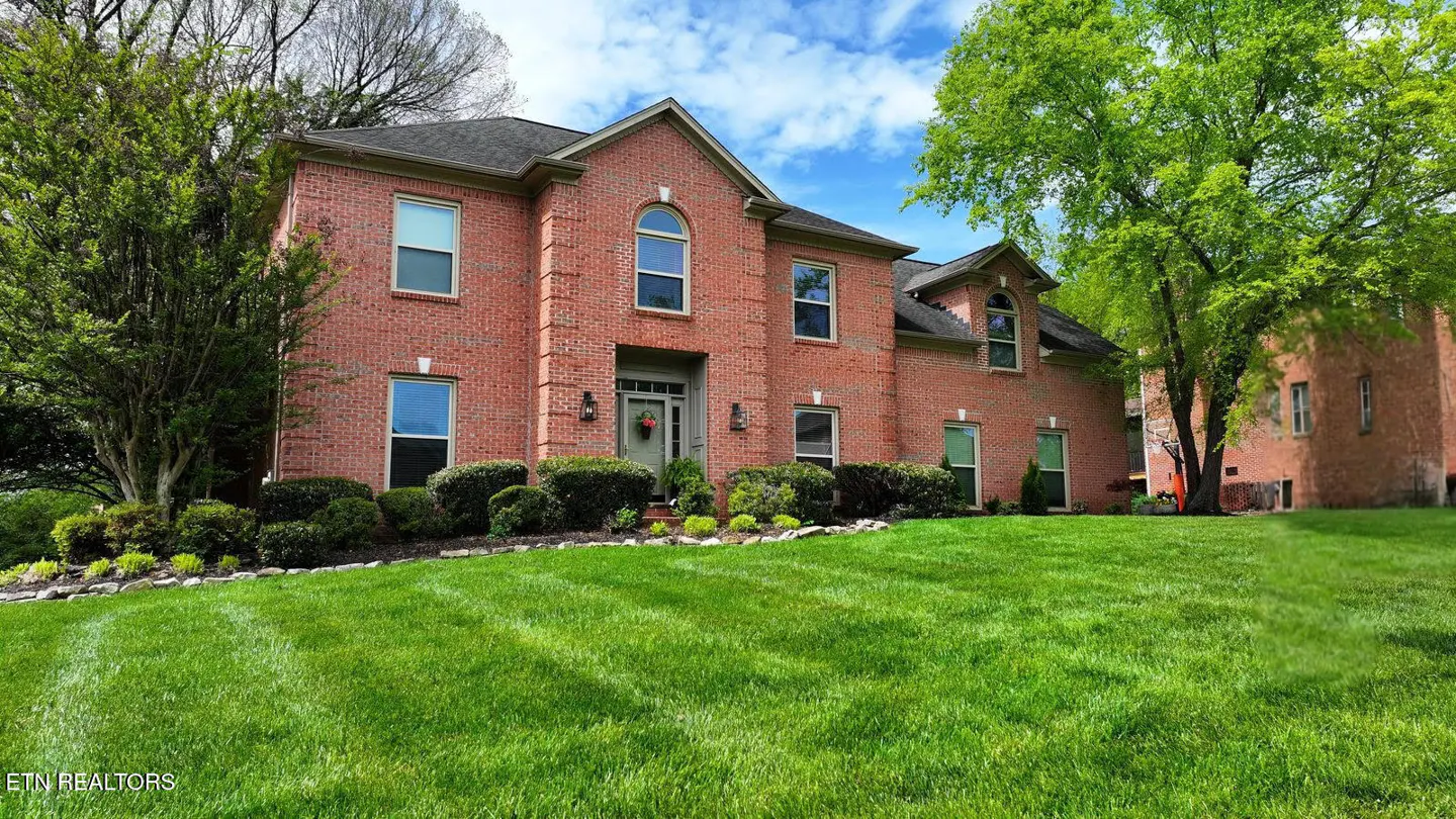 Two-story red brick house with a green lawn, bushes, and trees under a blue sky with white clouds.