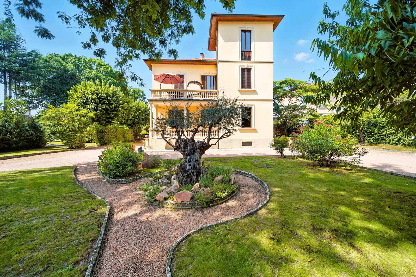 A three-story cream-colored house with a balcony and red umbrella, surrounded by green trees and a gravel path.