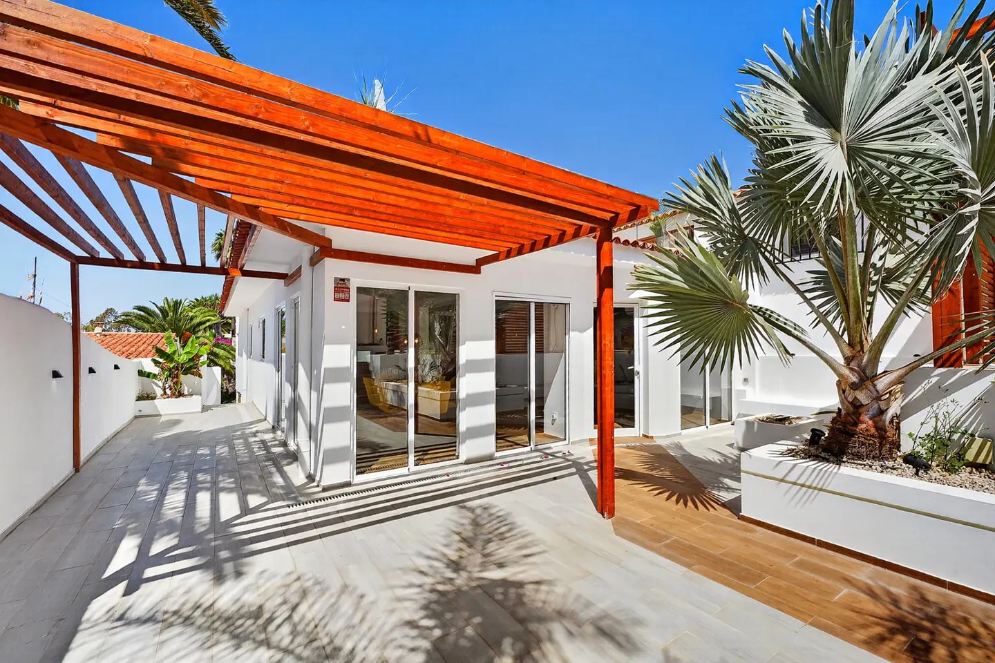 Sunny patio with a wooden pergola, white walls, glass sliding doors, and a large palm tree against a clear blue sky.