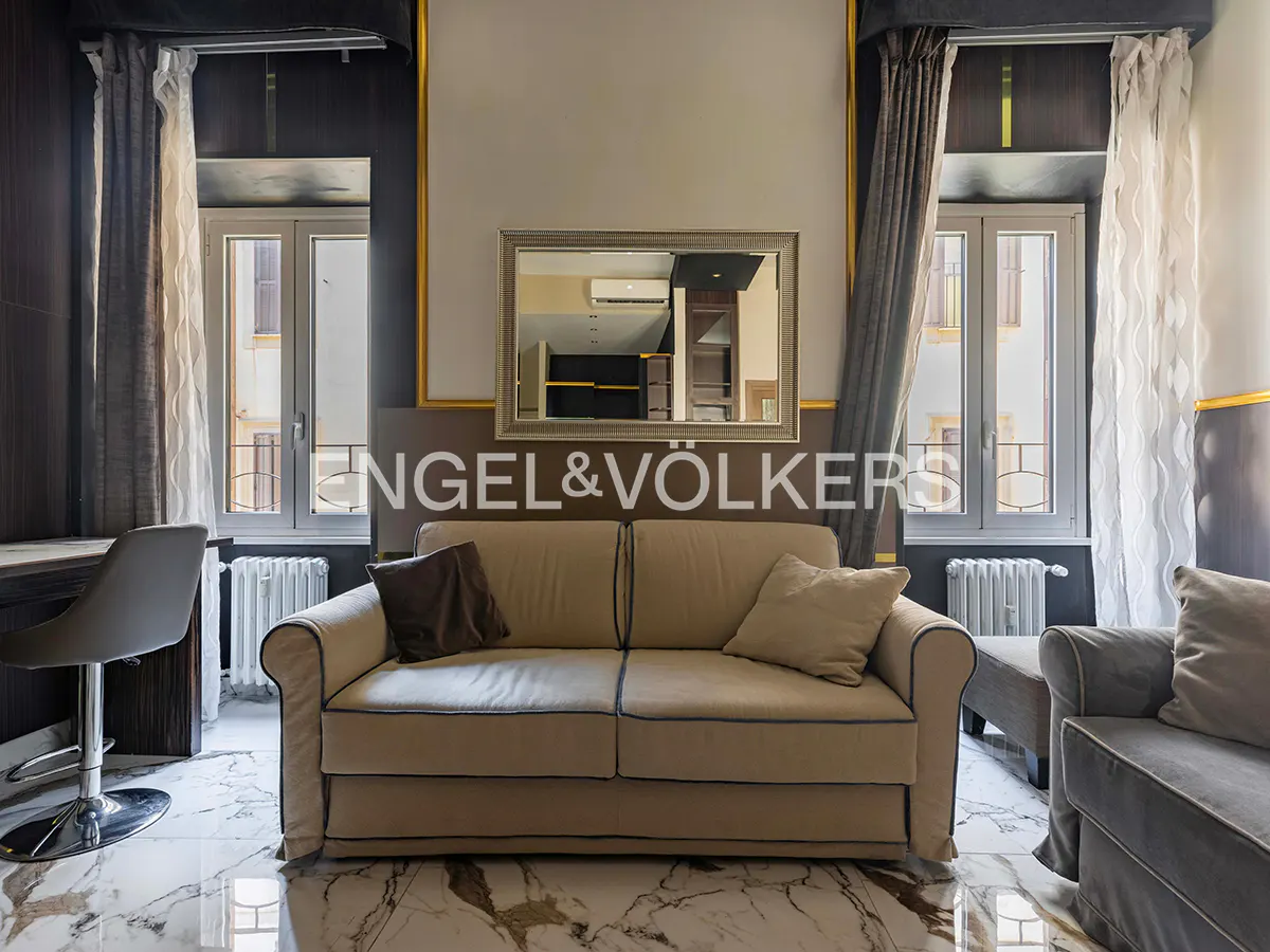 Living room with beige sofa, brown pillows, marble floor, desk, chair, and mirror reflecting another room. Windows with curtains on either side.