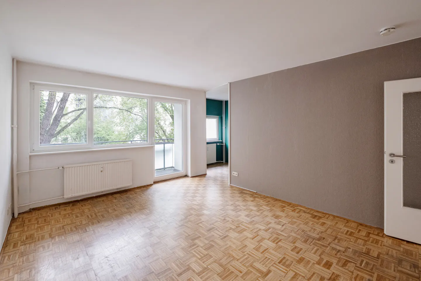 Bright, empty room with parquet floors, a large window, and a gray accent wall. A white door is on the right.