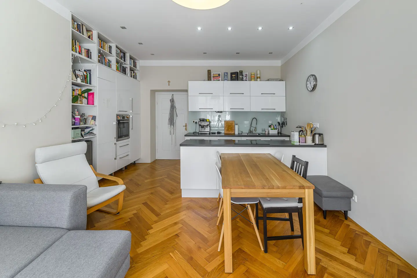 Bright, modern apartment interior with a wooden dining table, white kitchen cabinets, and a gray sofa on a herringbone wood floor.