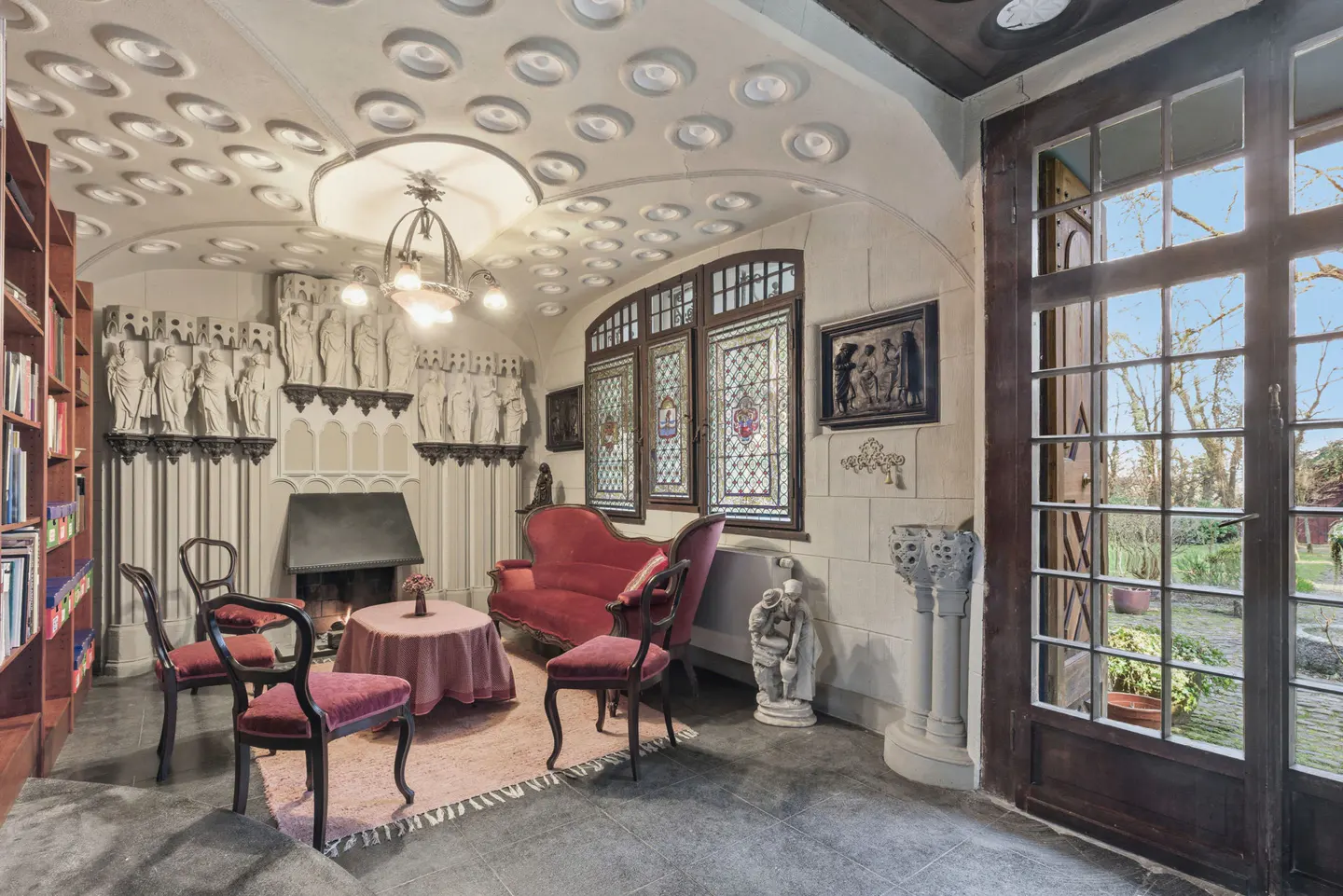 A cozy reading room with a fireplace, red furniture, and a wall of books. Stained glass windows and a unique ceiling add character.