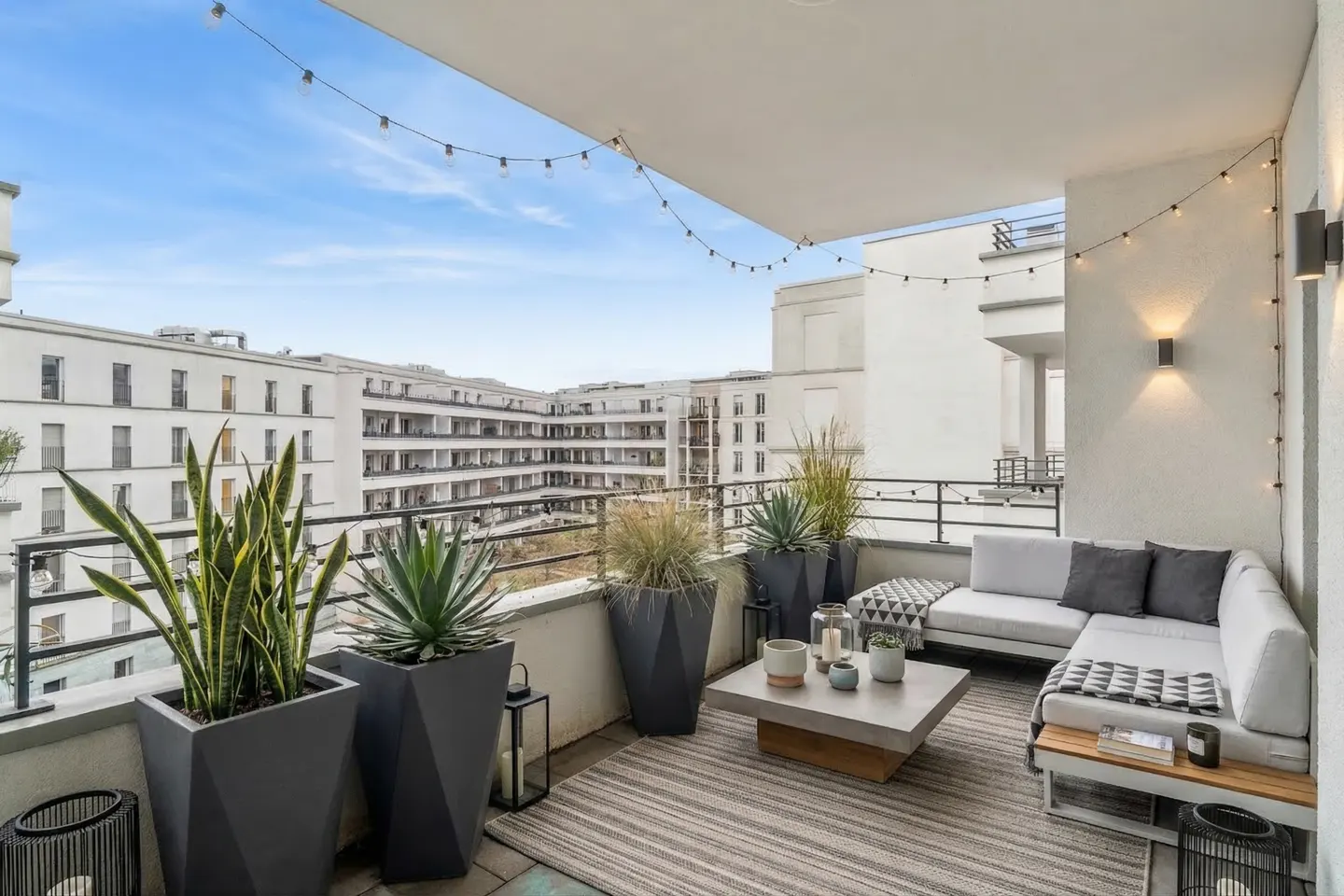 Cozy balcony with a gray sectional sofa, concrete table, and potted plants. String lights hang above, with city buildings in the background.