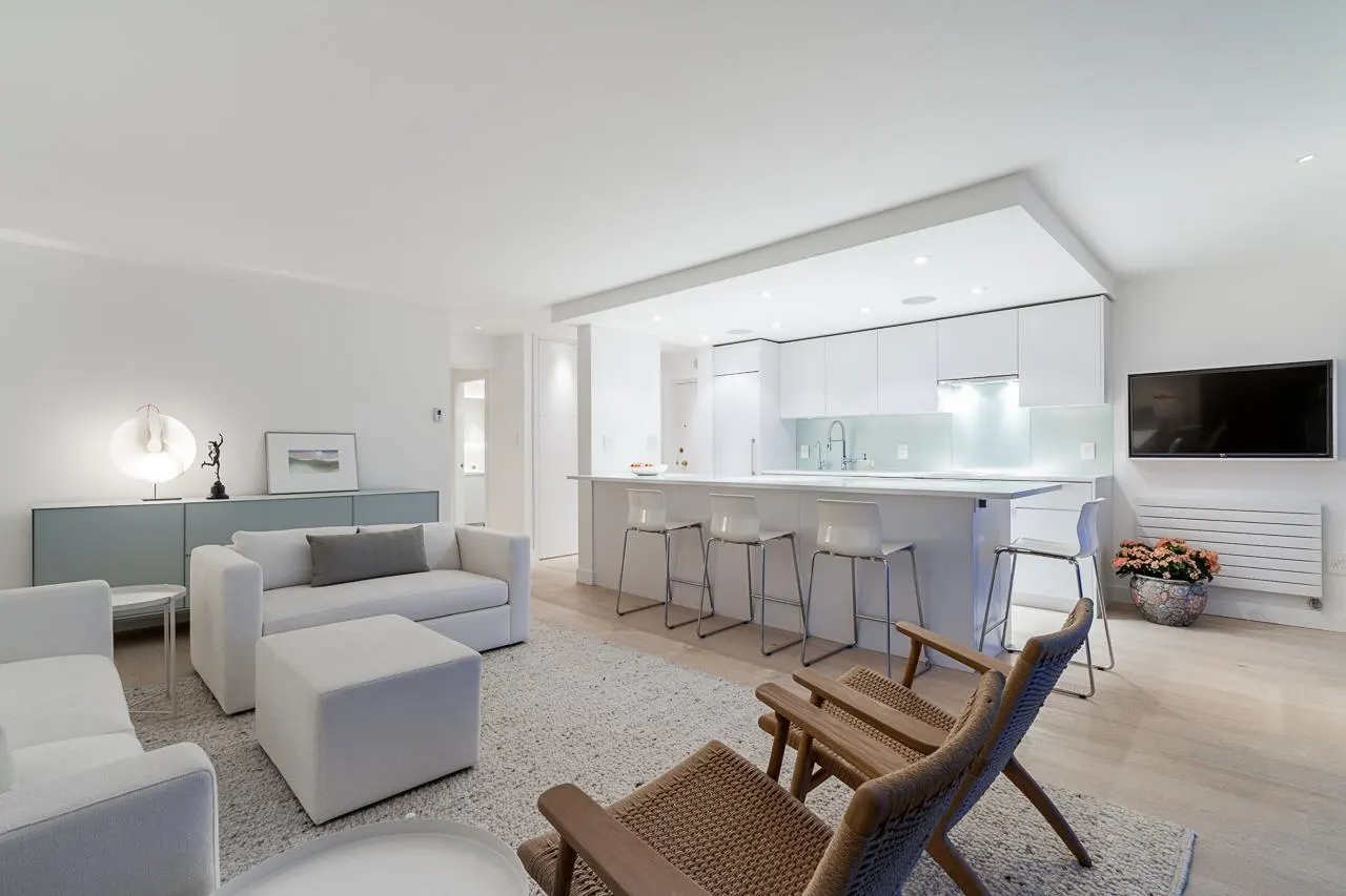 Bright, open-concept living space with white sofas, a kitchen island with stools, and wooden chairs on a textured rug. A TV hangs above a radiator.
