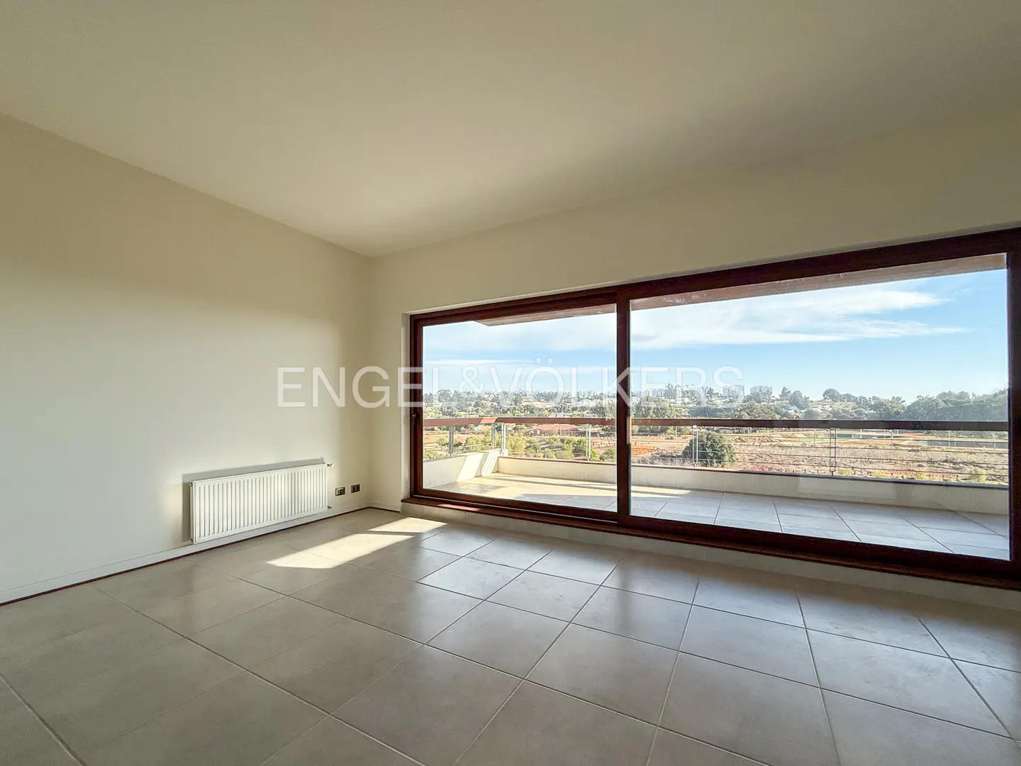 Bright, empty room with tile floor and large sliding glass doors to a balcony with a view. A white radiator is on the wall.