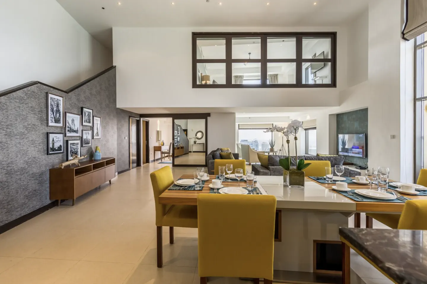 Bright, modern living room with yellow chairs around a set dining table. A staircase and gallery wall are on the left. A large window overlooks the city.