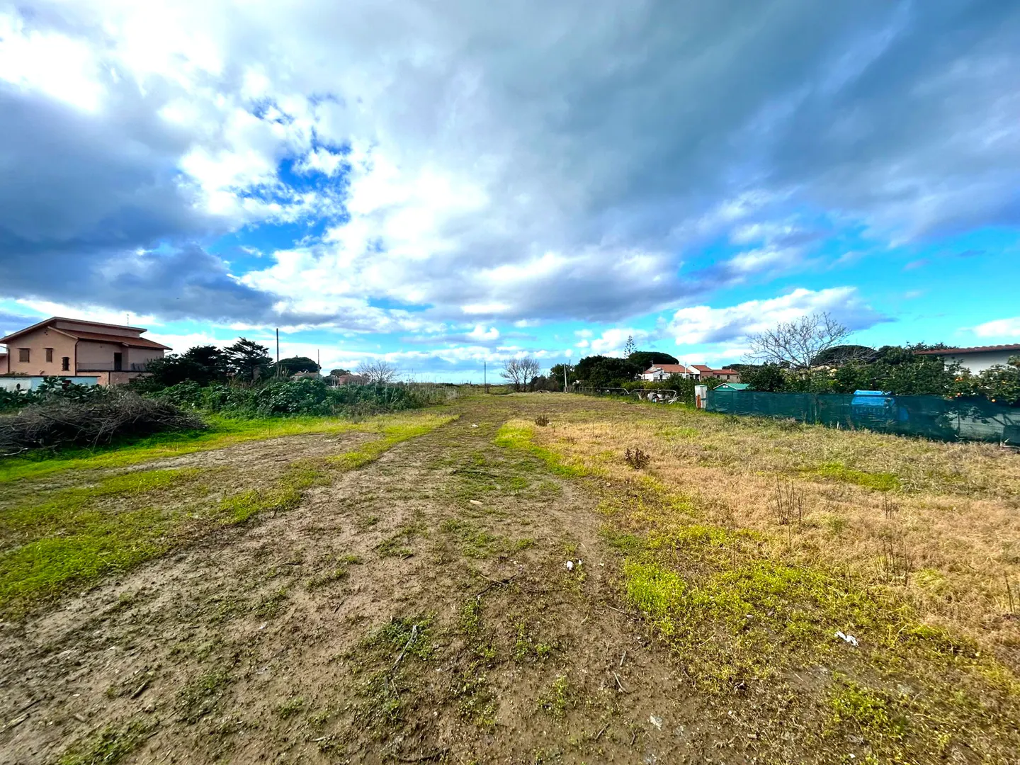 Vacant land with a dirt path, green and brown grass, and scattered trees under a cloudy blue sky. Houses are visible in the background.
