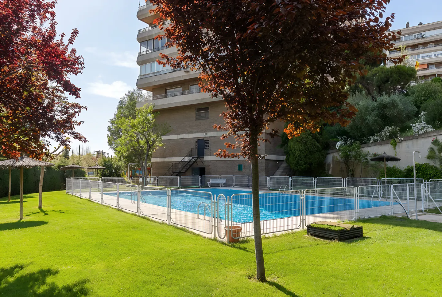 Outdoor pool with blue water, surrounded by a white fence and green grass. Trees and a building are in the background.