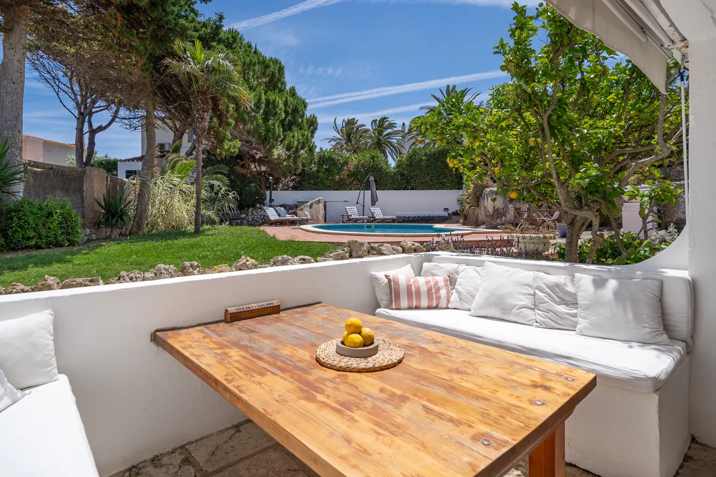 Covered patio with wooden table, bowl of lemons, and white cushioned seating overlooking a sunny garden and swimming pool.